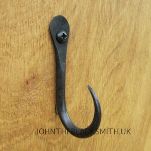 Könnte beinhalten: Ein schwarzer Metallhaken mit einem gebogenen Ende, montiert auf einer Holzoberfläche. Der Haken ist mit einer Schraube befestigt. Der Text "JOHNTHEBLACKSMITH.UK" ist unterhalb des Hakens sichtbar.