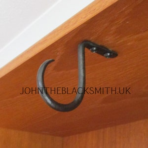 Może przedstawiać: Czarny metalowy hak z zakrzywionym końcem, zamontowany na drewnianej powierzchni. Hak jest przymocowany śrubami i ma prosty, funkcjonalny design. Tekst "JOHNTHEBLACKSMITH.UK" jest widoczny na zdjęciu.
