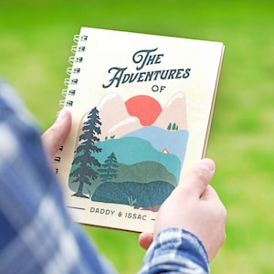May include: A wooden spiral bound notebook with a colorful illustration of mountains, a red sun, and a tent. The text on the cover reads 'The Adventures Of Daddy & Issac'.