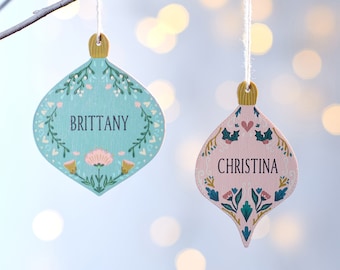 Può includere: Due addobbi natalizi personalizzati, uno azzurro chiaro con il nome "Brittany" e l'altro rosa con il nome "Christina". Entrambi gli addobbi presentano un design floreale.