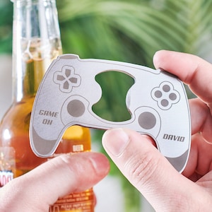 Può includere: Un apribottiglie argentato a forma di controller per videogiochi. L'apribottiglie ha le parole "Game On" e "David" stampate su di esso.