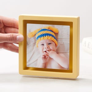 May include: A square, yellow picture frame holding a photograph of a baby wearing a blue and yellow knit hat. The baby is smiling and has their fingers in their mouth. The frame is sitting on a white surface.