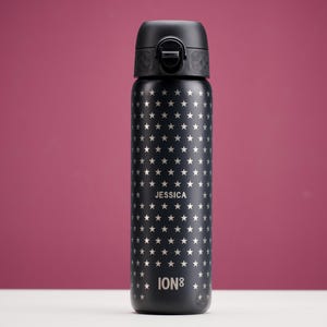 Puede incluir: Botella de agua aislada negra con un patrón de estrellas y una tapa negra. El nombre "JESSICA" está impreso en el centro, y la marca "ION8" está en la parte inferior. La botella está sobre una superficie blanca con un fondo burdeos.