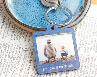 Puede incluir: Un llavero de madera azul con una foto de un padre y un hijo pescando. El llavero tiene el texto "Best Dad in the World" impreso en él.