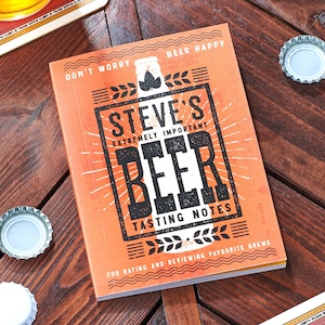 May include: An orange notebook with the text "STEVE'S EXTREMELY IMPORTANT BEER TASTING NOTES" in black. The notebook is open on a wooden table, with beer bottle caps scattered around. The text "DON'T WORRY BEER HAPPY" is at the top.