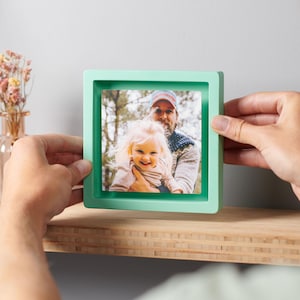 May include: A square, mint-green picture frame holding a color photograph of a smiling child and an adult. The frame is held by a person's hands, with a wooden shelf and a vase of dried flowers in the background.