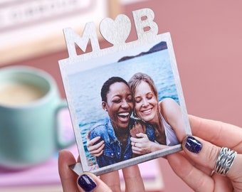 Puede incluir: Marco de fotos plateado con un corazón recortado y las letras "M" y "B" en la parte superior. El marco contiene una foto de dos mujeres sonriendo y abrazándose en una playa.