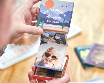 Puede incluir: Un álbum de fotos azul y naranja con una escena de montaña y el texto "Daddy & Amelie's Adventures Since 2018". El álbum está abierto para revelar fotos de una familia.