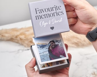 Puede incluir: Una caja gris con el texto "Favourite memories Olga & David" y un corazón. La caja está abierta y contiene una pila de fotos.