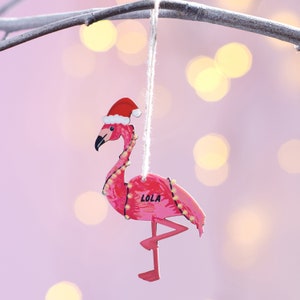 May include: Pink flamingo Christmas ornament with a Santa hat and the name "LOLA" written on the body.