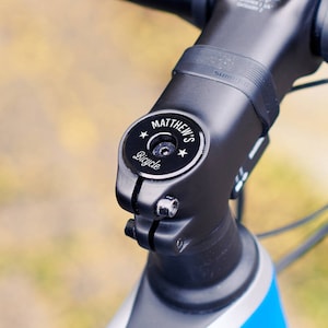 Close-up of a black personalised bike headset cap installed on the handlebar stem, engraved with “MATTHEW’S” and “Bicycle”, with star motifs.