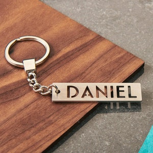 Può includere: Portachiavi in ​​argento con una catena in argento e un'etichetta rettangolare con il nome "DANIEL" ritagliato.