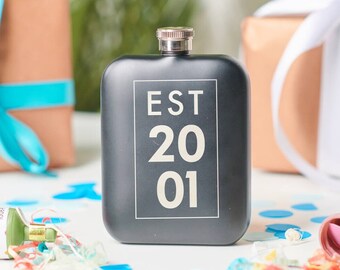 May include: Black hip flask with a silver top and the text "EST 2001" printed in white on a black rectangle.