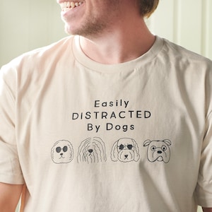 May include: A beige t-shirt featuring the phrase "Easily Distracted By Dogs" and four black cartoon dog illustrations. The dogs have varied appearances. The shirt is made of a soft material and is a neutral colour.
