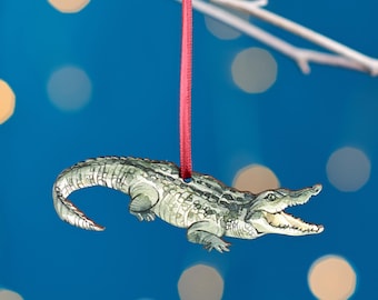 May include: A green and gray alligator Christmas ornament with a red ribbon hanging from the top. The alligator has its mouth open and its teeth are visible.