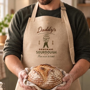 May include: A beige linen apron featuring a dark brown graphic print that reads Daddy's, 2026, HOMEMADE SOURDOUGH, and From oven to table, with an illustration of a person holding a loaf of bread above their head, worn over a dark green shirt.