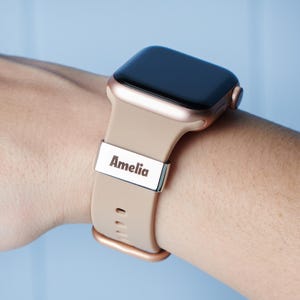 Op de afbeelding: Een Apple Watch in rosé goud met een beige siliconen band. De band heeft een zilveren gepersonaliseerde plaat met de naam "Amelia" erop gegraveerd.