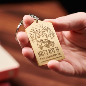 May include: A gold-coloured metal keychain with a laser-engraved design. The design features a mountain landscape with a Land Rover vehicle and the text "Matt's Keys to The Landrover".