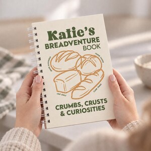 Könnte beinhalten: Ein spiralgebundenes Notizbuch mit cremefarbenem Einband, das den grünen Text Katie's BREADVENTURE BOOK und CRUMBS, CRUSTS & CURIOSITIES zeigt. Der Einband enthält braune Strichzeichnungen verschiedener Brotlaibe mit kleinen Textbeschriftungen.