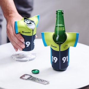Könnte beinhalten: Eine grüne Bierflasche und eine Dose in passenden marineblauen und gelben Trikot-Stil-Kühlern. Die Kühler haben die Nummer "1991" und den Namen "Thomas" aufgedruckt. Ein Flaschenöffner und ein Kronkorken liegen ebenfalls auf der weißen Oberfläche.