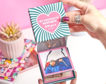 Puede incluir: Una caja a rayas azul turquesa y blanca con un corazón rosa que dice "Mis recuerdos favoritos contigo x Est. 2017". La caja está abierta y tiene una pila de fotos dentro.