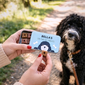 Puede incluir: Una lata de metal azul claro con la inscripción "BELLA'S ON THE GO TREATS", una ilustración de perro y gráficos de huesos. La lata contiene golosinas cuadradas para perros. Una mano sostiene una golosina, con un perro negro en el fondo.