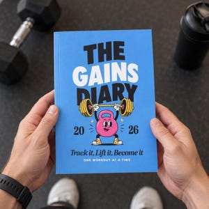Pode incluir: Um caderno azul intitulado THE GAINS DIARY 2026 com uma ilustração de um kettlebell rosa levantando uma barra. A capa apresenta o texto Track it, Lift it, Become it e ONE WORKOUT AT A TIME em letras pretas e brancas sobre o fundo azul.