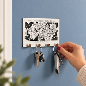 May include: A rectangular wall-mounted key holder featuring a black and white street map print. The text reads NO.24 WOODBURY PARK ROAD and THE CAMPBELL FAMILY. It includes four small metal hooks for hanging keys, with two sets of keys attached.