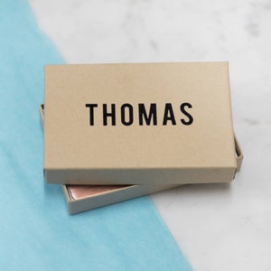 K&ouml;nnte beinhalten: Eine braune Pappbox mit dem Namen "THOMAS" in schwarzen Buchstaben auf der Oberseite.