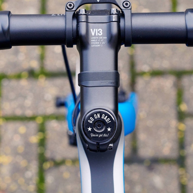 Black personalised bicycle headset cap fitted to the stem of a blue bike, engraved with “GO ON DAD!” and “You’ve got this!” alongside two stars.