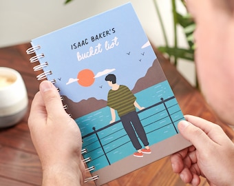 May include: A blue spiral bound notebook with the text "ISAAC BAKER'S bucket list" on the cover. The cover features an illustration of a person standing on a pier overlooking a body of water with a sunset in the background.