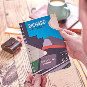 Op de afbeelding: Een blauw en grijs spiraalgebonden notitieboek met de tekst "Richard Peter's Train Spotting Journal" en een gestileerde illustratie van een trein op de omslag.