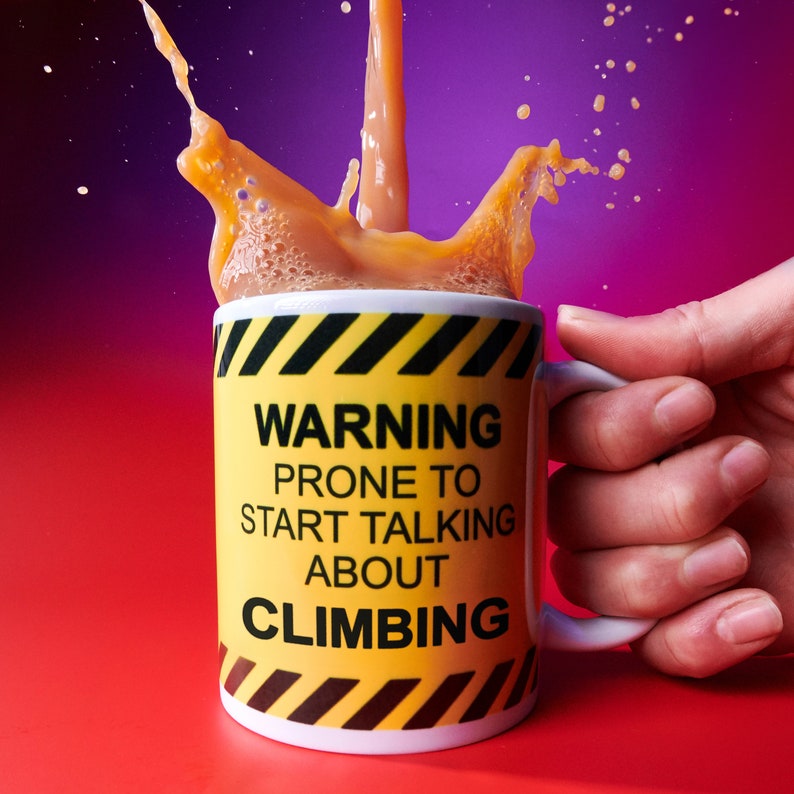 May include: A white mug with a yellow and black warning sign design. The mug reads "Warning Prone to Start Talking About Climbing".  The mug is filled with a brown liquid and is being held by a hand.