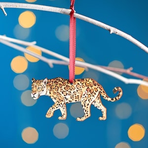 May include: A wooden ornament shaped like a jaguar with brown and black spots. The ornament is hanging from a red ribbon on a branch.