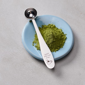 Puede incluir: Una cuchara medidora de metal plateado sobre un plato de cerámica azul claro que contiene un montón de polvo de matcha verde. El mango de la cuchara tiene grabado el texto "Isobel's little scoop of zen" y un pequeño gráfico de una taza.