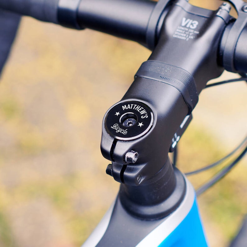 Close-up of a black personalised bike headset cap installed on the handlebar stem, engraved with “MATTHEW’S” and “Bicycle”, with star motifs.