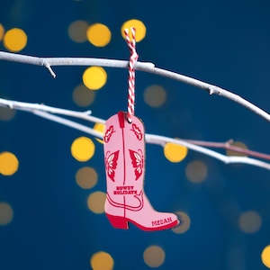 Puede incluir: Adorno de Navidad de bota vaquera rosa con el texto "Howdy Holidays" y el nombre "Megan" en la parte inferior.