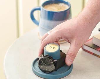 May include: A teal watch stand with a black watch and a black leather band. The stand has a cylindrical post with the initials "H.C." on a gold-colored top. A blue and white mug of coffee is in the background.