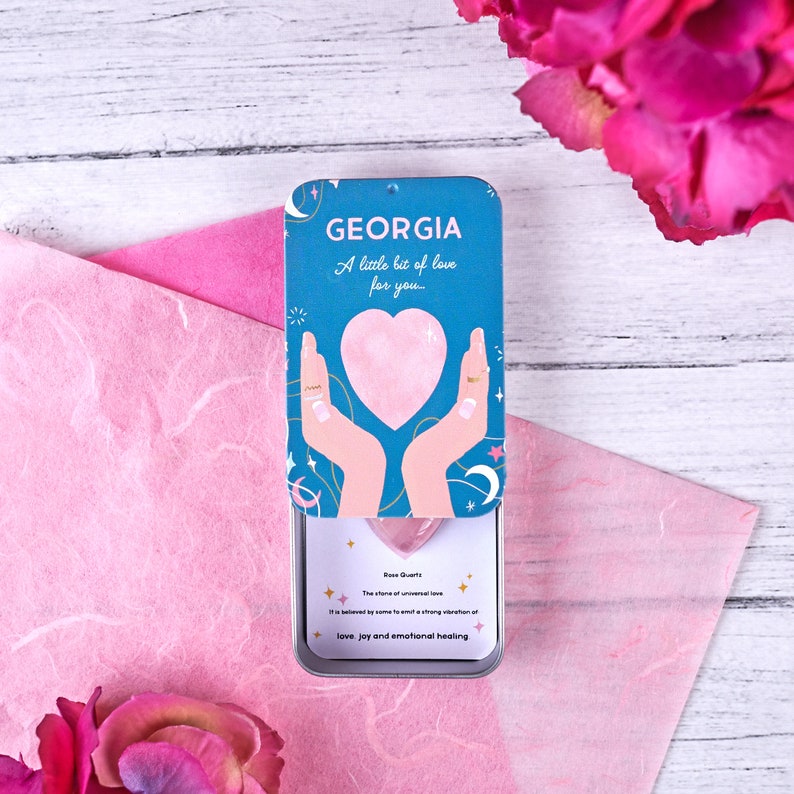 Open personalised “GEORGIA” crystal tin with a blue lid showing hands holding a pink heart and a rose quartz heart crystal with a message card inside.