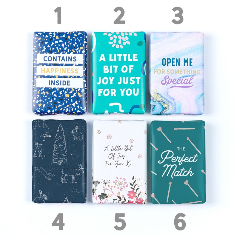 K&ouml;nnte beinhalten: Sechs verschiedene Seifenst&uuml;cke mit unterschiedlichen Designs und Texten. Die Seifenst&uuml;cke sind in zwei Reihen zu je drei angeordnet. Die obere Reihe zeigt ein blaues und wei&szlig;es Seifenst&uuml;ck mit dem Text "CONTAINS HAPPINESS INSIDE", ein t&uuml;rkisfarbenes Seifenst&uuml;ck mit dem Text "A LITTLE BIT OF JOY JUST FOR YOU" und ein lila und wei&szlig;es Seifenst&uuml;ck mit dem Text "OPEN ME FOR SOMETHING Special". Die untere Reihe zeigt ein dunkelblaues Seifenst&uuml;ck mit einer Waldszene, ein wei&szlig;es Seifenst&uuml;ck mit einem Blumendesign und dem Text "A Little Bit Of Joy For You X" und ein t&uuml;rkisfarbenes Seifenst&uuml;ck mit dem Text "THE Perfect Match".