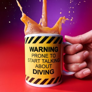 May include: A white ceramic mug with a yellow and black warning sign design. The mug has the text "WARNING PRONE TO START TALKING ABOUT DIVING" printed on it. The mug is filled with a brown liquid and is being poured into the mug, creating a splash.