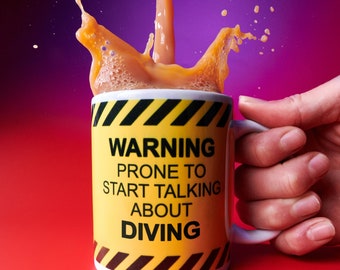 Puede incluir: Una taza de cerámica blanca con un diseño de señal de advertencia amarilla y negra. La taza tiene el texto "WARNING PRONE TO START TALKING ABOUT DIVING" impreso en ella. La taza está llena de un líquido marrón y se está vertiendo líquido en la taza, creando salpicaduras.