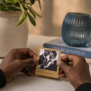 May include: A small, square photograph held by hands, showing a black and white image of two men in formal wear, embracing. The photograph has text that reads "Two souls, one beautiful adventure."