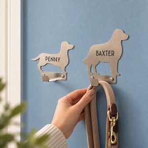 May include: Two brushed metal wall hooks shaped like dog silhouettes. The smaller hook is labeled with the name PENNY, and the larger hook is labeled with the name BAXTER. Each hook features a small metal shelf at the base for hanging items.