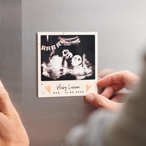 May include: A Polaroid-style photograph of a baby scan, displaying a black and white image of a baby. The photo has the text "Baby Larson" and "DUE - 14.09.2025". Decorated with small hearts and stars.