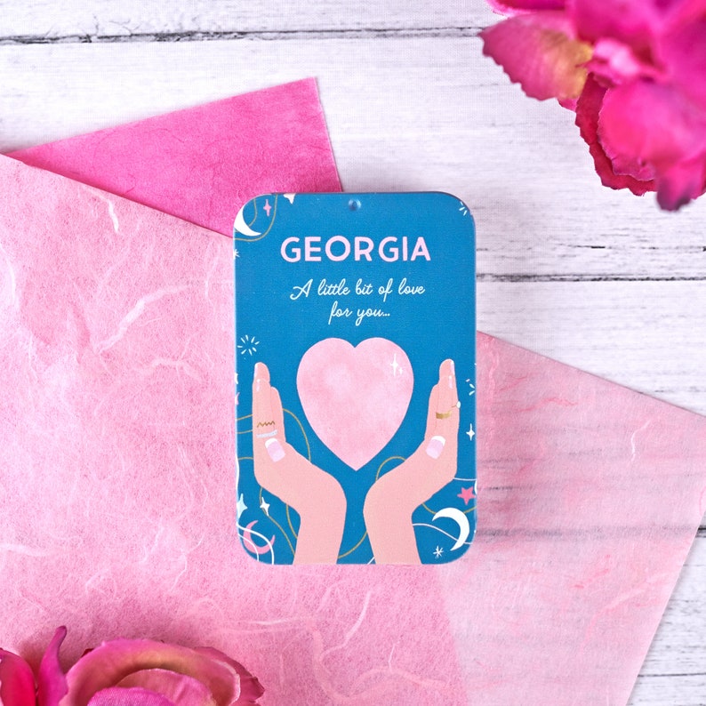 Blue personalised aluminium pocket tin labelled “GEORGIA” with “A little bit of love for you...” and an illustration of hands holding a pink heart, stars and moons.