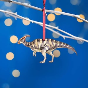 Watercolour Parasaurolophus wooden Christmas tree decoration hanging from a burgundy ribbon against blue bokeh lights, showing its long crest, striped body and tail.