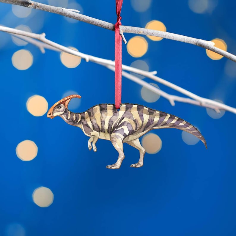 Watercolour Parasaurolophus wooden Christmas tree decoration hanging from a burgundy ribbon against blue bokeh lights, showing its long crest, striped body and tail.