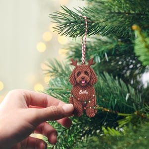 May include: A brown dog-shaped Christmas ornament featuring reindeer antlers and a string of colorful holiday lights. The name Bella is printed in white script across the chest of the dog. The ornament hangs from a red and white twine string.