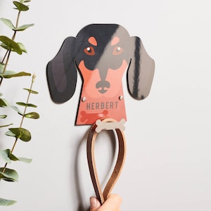 Może przedstawiać: Hak ścienny w kształcie głowy psa rasy dachshund w kolorze czerwonym i czarnym z nadrukowanym napisem "Herbert". Do haka przymocowany jest brązowy skórzany smycz.